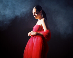 A beautiful girl with a long ponytail stands in a red gown with nothing but smoke clouds and a spotlight around her.