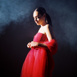 A beautiful girl with a long ponytail stands in a red gown with nothing but smoke clouds and a spotlight around her.