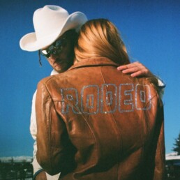 Urban cowboy gently pulling back the hair of a back-turned RODEO queen in a be-dazzled leather jacket.