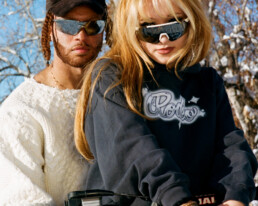 Male and female models straddle motocross bike wearing custom RODEO the Label winter 2022 capsule collection items and reflective sunglasses.