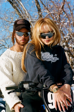 Male and female models straddle motocross bike wearing custom RODEO the Label winter 2022 capsule collection items and reflective sunglasses.
